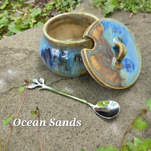 May include: A ceramic jar with a lid, featuring blue and brown glaze, and a silver spoon with a leaf design. The jar is open, revealing its interior. The text "Ocean Sands" is visible.