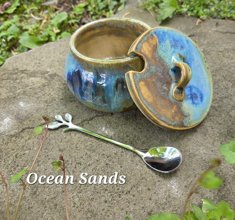 May include: A ceramic jar with a lid, featuring blue and brown glaze, and a silver spoon with a leaf design. The jar is open, revealing its interior. The text "Ocean Sands" is visible.