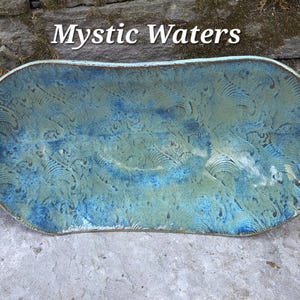 May include: A handmade ceramic platter with a unique, organic shape. The platter features a blend of blue and green hues, with a textured surface and a decorative pattern. The words "Mystic Waters" are displayed in white text.