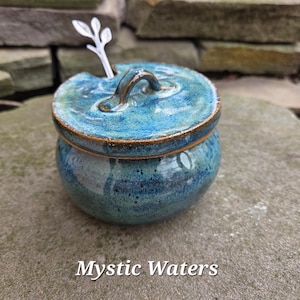 May include: A handcrafted ceramic jar with a lid and a small spoon. The jar is a deep blue-green color with a speckled glaze and a bronze-colored rim. The lid has a handle and the text "Mystic Waters" is at the bottom.