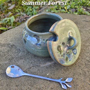 May include: A ceramic jar with a lid and a stainless steel spoon. The jar is glazed in shades of blue and green. The lid has a handle and a matching glaze. The spoon has a leaf design at the end. The text "Summer Forest" is at the top.