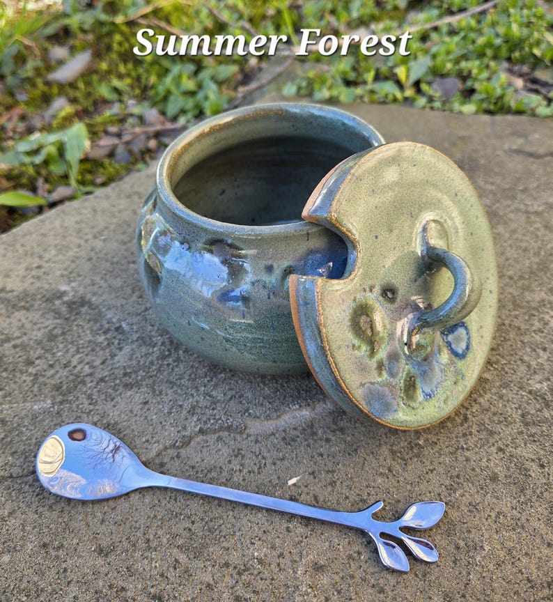 May include: A ceramic jar with a lid and a stainless steel spoon. The jar is glazed in shades of blue and green. The lid has a handle and a matching glaze. The spoon has a leaf design at the end. The text "Summer Forest" is at the top.