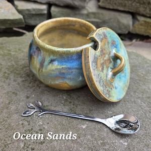 May include: A ceramic jar with a lid, featuring blue, green, and gold tones. A small, silver spoon with a leaf design rests beside the jar. The text "Ocean Sands" is displayed at the bottom of the image.
