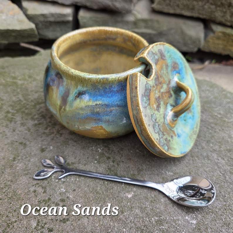 May include: A ceramic jar with a lid, featuring blue, green, and gold tones. A small, silver spoon with a leaf design rests beside the jar. The text "Ocean Sands" is displayed at the bottom of the image.