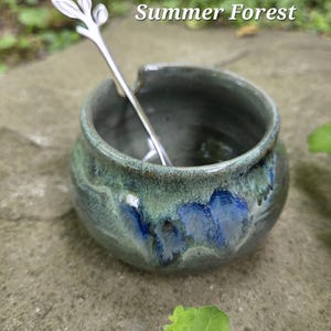 May include: A small, handmade ceramic bowl with a green and blue glaze, holding a silver spoon. The spoon has a leaf-like design at the top. The text "Summer Forest" is above the bowl.