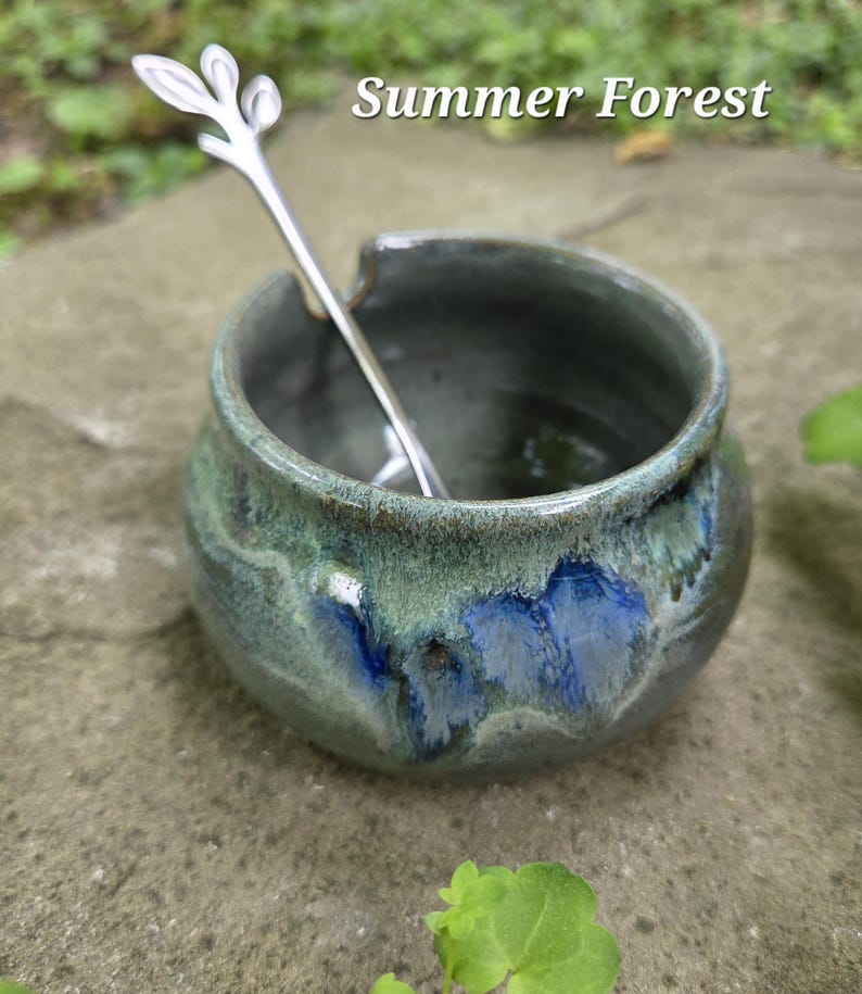 May include: A small, handmade ceramic bowl with a green and blue glaze, holding a silver spoon. The spoon has a leaf-like design at the top. The text "Summer Forest" is above the bowl.