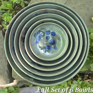 May include: A set of six nested ceramic bowls in a sage green glaze with dark blue and brown speckled accents. The bowls are arranged in a circular stack, showing a smooth interior finish and a consistent color palette throughout the set.