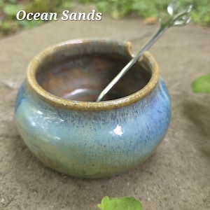 May include: A small, round ceramic bowl with a blue and green glaze and a brown rim. A small, silver spoon with a leaf-shaped handle rests inside the bowl. The text "Ocean Sands" is above the bowl.