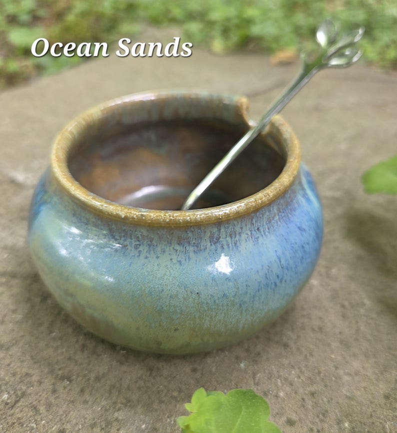 May include: A small, round ceramic bowl with a blue and green glaze and a brown rim. A small, silver spoon with a leaf-shaped handle rests inside the bowl. The text "Ocean Sands" is above the bowl.
