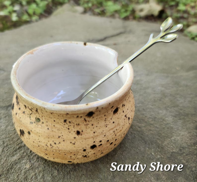 May include: A speckled ceramic bowl with a silver spoon. The bowl is tan with dark brown spots and a white interior. The spoon has a leaf-like design on the handle. The text "Sandy Shore" is at the bottom.