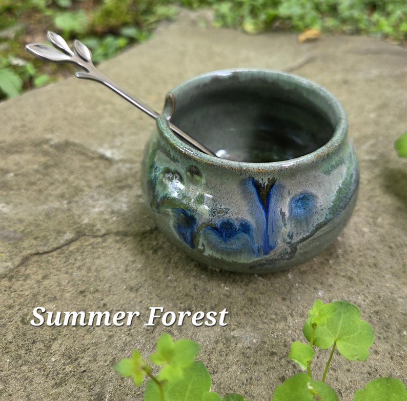 May include: A small, round ceramic bowl with a green and blue glaze, and a silver spoon with a leaf design. The bowl is sitting on a stone surface. The text "Summer Forest" is at the bottom of the image.