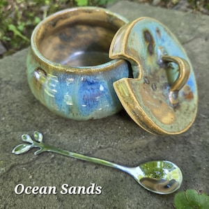 May include: A round ceramic jar with a lid featuring a glossy glaze in shades of blue, green, and brown, accompanied by a silver-toned metal spoon with a decorative leaf-shaped handle. The text "Ocean Sands" is printed in white at the bottom.