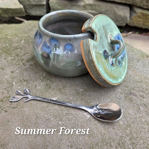 May include: A ceramic jar with a lid, glazed in shades of green, blue, and gray, with a matching spoon. The lid is partially open, revealing the interior. The spoon has a leaf-like design on the handle. Text on the image reads "Summer Forest."