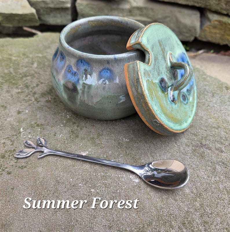 May include: A ceramic jar with a lid, glazed in shades of green, blue, and gray, with a matching spoon. The lid is partially open, revealing the interior. The spoon has a leaf-like design on the handle. Text on the image reads "Summer Forest."