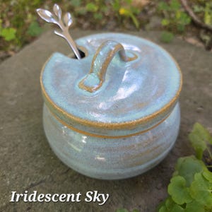 May include: A light blue ceramic sugar bowl with a matching lid and a silver-toned metal spoon featuring a leaf-shaped handle. The bowl has a glossy finish with visible brown clay edges and the text Iridescent Sky printed at the bottom.
