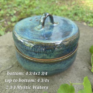 May include: A handcrafted ceramic container with a lid. The container is a blend of blue and green hues, with a glossy finish. It features a curved handle on top and a circular shape. The text on the container reads: "bottom: 4 3/4x1 3/4, top to bottom: 4 3/4x, 2 3/4 Mystic Waters."