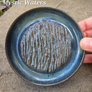 May include: A round, ceramic dish with a textured center and a glossy, blue glaze. The center features a raised, linear pattern in shades of brown and teal. The dish is labeled "Mystic Waters" in white text.