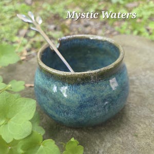May include: A small, handcrafted ceramic bowl with a teal and blue glaze, featuring a dark green rim. A decorative silver spoon rests inside the bowl. The text "Mystic Waters" is displayed in white letters above the bowl.