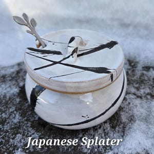 May include: A white ceramic sugar bowl with a lid and a small handle. The bowl is decorated with black paint splatters. A small silver spoon rests in a slot on the lid. The text "Japanese Splatter" is at the bottom.