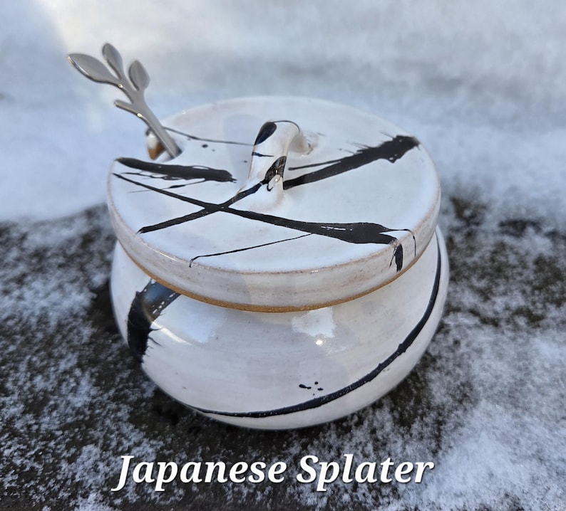 May include: A white ceramic sugar bowl with a lid and a small handle. The bowl is decorated with black paint splatters. A small silver spoon rests in a slot on the lid. The text "Japanese Splatter" is at the bottom.