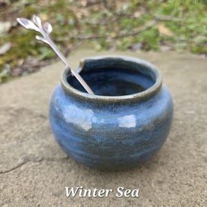 May include: A small, round, blue ceramic jar with a silver spoon inside. The jar has a dark blue rim and a glossy finish. The text "Winter Sea" is at the bottom of the image.