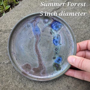 May include: A round ceramic plate with a 5 inch diameter, featuring a mottled gray and brown glaze with vibrant blue crystalline patterns. The text Summer Forest and 5 inch diameter is printed on the surface of the plate.