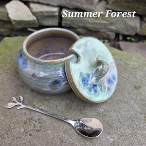 May include: A ceramic sugar bowl with a lid, featuring a green and blue glaze. The lid has a handle and floral accents. A silver spoon with a leaf design rests beside the bowl. The text "Summer Forest" is visible.