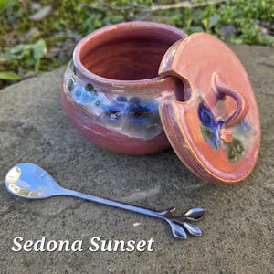 May include: A ceramic jar with a lid, in shades of red, blue, and orange, with a matching lid and a small silver spoon. The jar has a round shape with a lid that has a handle. The spoon has a leaf design at the end. The text "Sedona Sunset" is at the bottom.