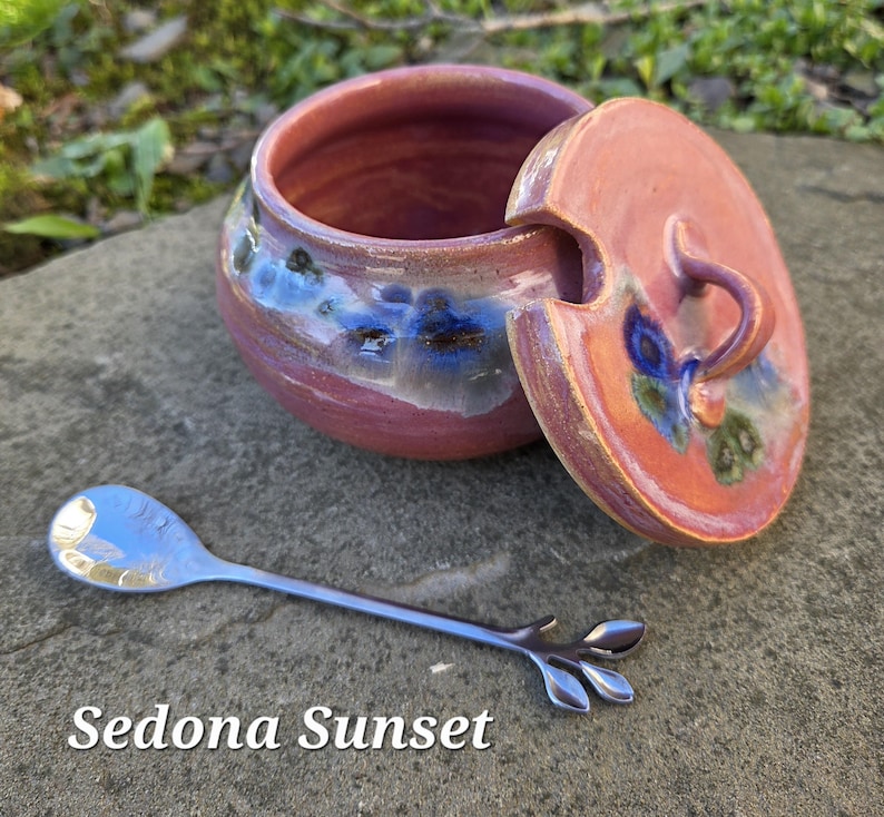 May include: A ceramic jar with a lid, in shades of red, blue, and orange, with a matching lid and a small silver spoon. The jar has a round shape with a lid that has a handle. The spoon has a leaf design at the end. The text "Sedona Sunset" is at the bottom.