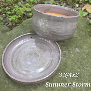 May include: A ceramic bowl and matching saucer set featuring a glossy, variegated lavender and grey glaze with subtle pink undertones. The set is labeled 3 3/4x2 Summer Storm, indicating dimensions of 3.75 inches by 2 inches for the bowl.