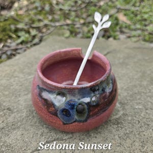 May include: A small, round, ceramic bowl in shades of red and brown, with a silver spoon resting inside. The spoon has a leaf design at the top. The text "Sedona Sunset" is at the bottom of the image.