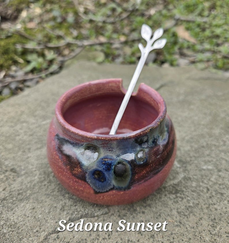 May include: A small, round, ceramic bowl in shades of red and brown, with a silver spoon resting inside. The spoon has a leaf design at the top. The text "Sedona Sunset" is at the bottom of the image.