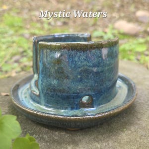 May include: A handcrafted ceramic sponge holder with a deep teal glaze. The holder has a cylindrical shape with a built-in dish and drainage hole. The text "Mystic Waters" is visible at the top. This unique piece adds a touch of artistry to any kitchen.
