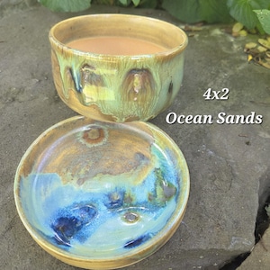 May include: A ceramic bowl and matching saucer set featuring a reactive glaze in shades of ocean blue, sandy tan, and seafoam green. The text 4x2 Ocean Sands is printed on the right side. The bowl has a smooth, rounded rim and a deep interior.