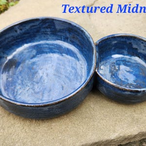 May include: A double-bowl ceramic dish with a deep blue glaze and a textured finish. The piece features one large circular bowl joined to a smaller circular bowl, with the text Textured Midnight displayed in blue above the object.