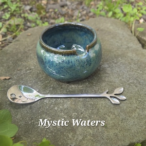 May include: A small, round, blue and green ceramic bowl with a brown rim and a stainless steel spoon with a leaf design. The text "Mystic Waters" is at the bottom of the image.