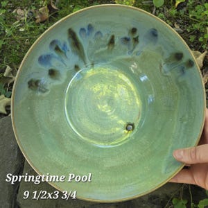 May include: A round ceramic bowl with a light green base glaze, featuring a darker green and blue drip pattern around the inner rim. The bowl is labeled Springtime Pool and measures 9 1/2 by 3 3/4 inches as indicated by the text on the image.