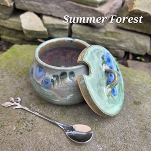 May include: A ceramic jar with a lid, featuring blue floral designs and a green and brown color scheme. A silver spoon with a leaf design rests beside the jar. The text "Summer Forest" is visible.