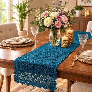 May include: A teal blue crochet table runner with a scalloped edge and intricate openwork pattern, centered on a wooden dining table. The runner is decorated with a glass vase of mixed flowers and two gold-toned candle holders with white candles.