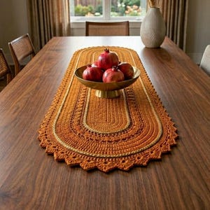 May include: An oval-shaped, burnt orange crocheted table runner with a scalloped edge and a central gold-toned braided accent. A gold-toned bowl sits in the center of the runner, containing four red pomegranates, placed on a wooden table.