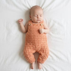Może przedstawiać: Brzoskwiniowy, ręcznie robiony na drutach pajacyk niemowlęcy o fakturze wafla, z kwadratowym dekoltem i dwoma jasnymi drewnianymi guzikami na ramiączkach. Ubranko posiada ściągacze przy kostkach i charakteryzuje się bezrękawowym krojem.