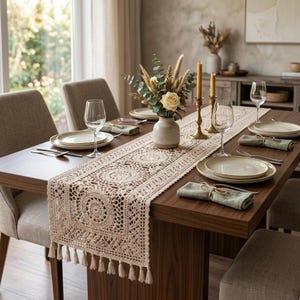 May include: A beige crochet table runner with intricate circular patterns and fringe tassels on the ends. The runner is placed on a dark wood dining table set with white ceramic plates, clear wine glasses, and sage green cloth napkins.