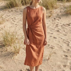 May include: A sleeveless midi-length dress in a solid terracotta color. The garment features a round neckline and a side-tie detail at the waist that creates a draped, wrap-style effect across the front of the skirt. The fabric has a textured finish.