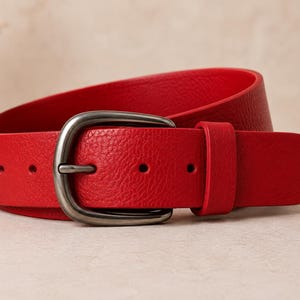 May include: A bright red belt with a textured surface and a rounded gunmetal gray metal buckle. The belt features a single matching red leather loop and a series of punched holes for adjustment, showcasing a simple and functional design.