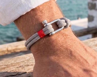 May include: A bracelet featuring a light gray cord wrapped in a double strand, secured by a polished silver-toned metal shackle clasp. A single vibrant red thread accent is wrapped around one section of the gray cord near the metal hardware.