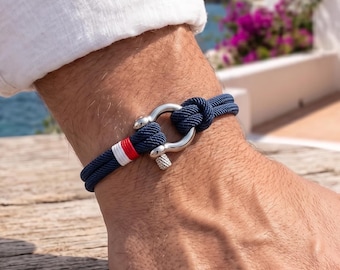 May include: A navy blue rope bracelet featuring a silver-toned metal shackle clasp. The cord is accented with a small white and red striped detail near the closure. The bracelet is constructed from a double-strand nautical style cord.