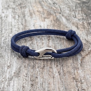 May include: Navy blue rope bracelet with a silver-colored clasp. The bracelet is made of two strands of rope, knotted at each end. The clasp is a curved, hook-like design. The bracelet is displayed on a weathered wooden surface.