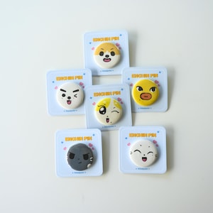 May include: Six circular pin-back buttons, each mounted on a square white backing card labeled ENCHIN PIN. The buttons feature various cartoon character faces with expressions including winking, smiling, and neutral, in colors of yellow, white, and gray.