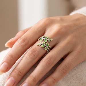 Puede incluir: Un anillo abierto de tono plateado que presenta dos ramas superpuestas con pequeñas hojas detalladas. Cada hoja está acentuada con un relleno de esmalte verde vibrante, creando un diseño inspirado en la naturaleza que envuelve el dedo con un aspecto delicado.