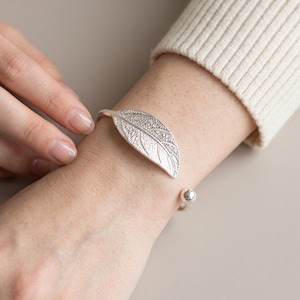 May include: A silver-toned cuff bracelet featuring a textured leaf design on one end and a small polished spherical bead on the other. The metal surface of the leaf shows detailed vein patterns, and the open-ended band is worn on the wrist.